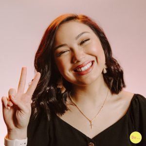 Woman with short brunette hair smiling holding a peace with her fingers.